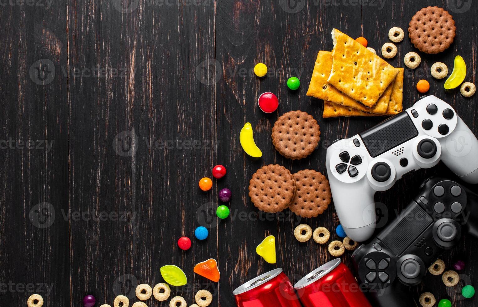 black and white modern gamepads on a dark wooden background among sweets, biscuits and cans of soda. photo