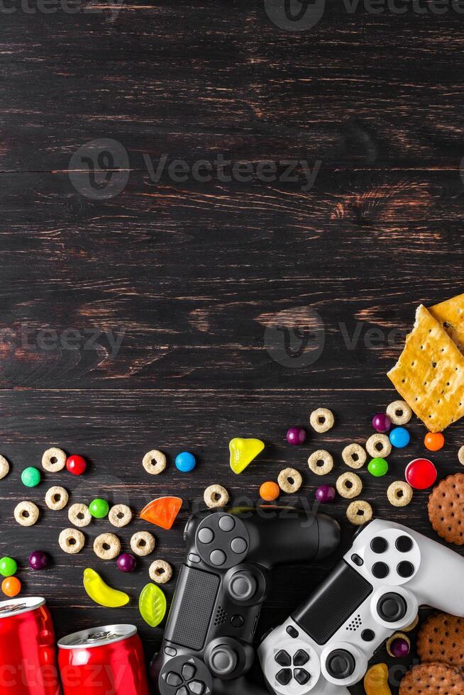 black and white modern gamepads on a dark wooden background among sweets, biscuits and cans of soda. photo