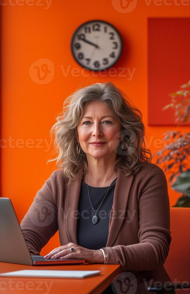 A smiling middle aged woman in an orange office, seated at her desk with a laptop photo