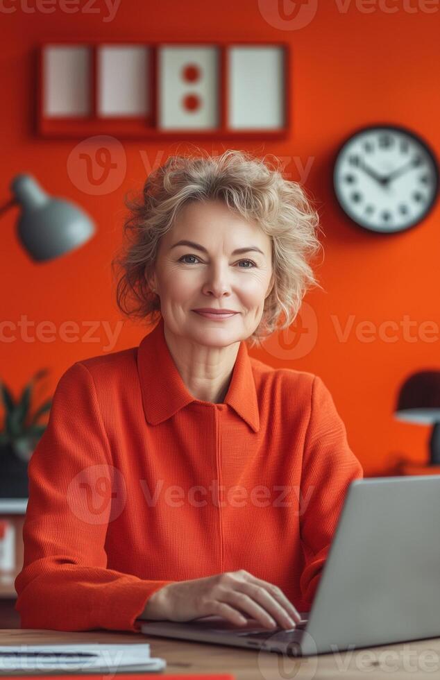 A smiling middle aged woman in an orange office, seated at her desk with a laptop photo