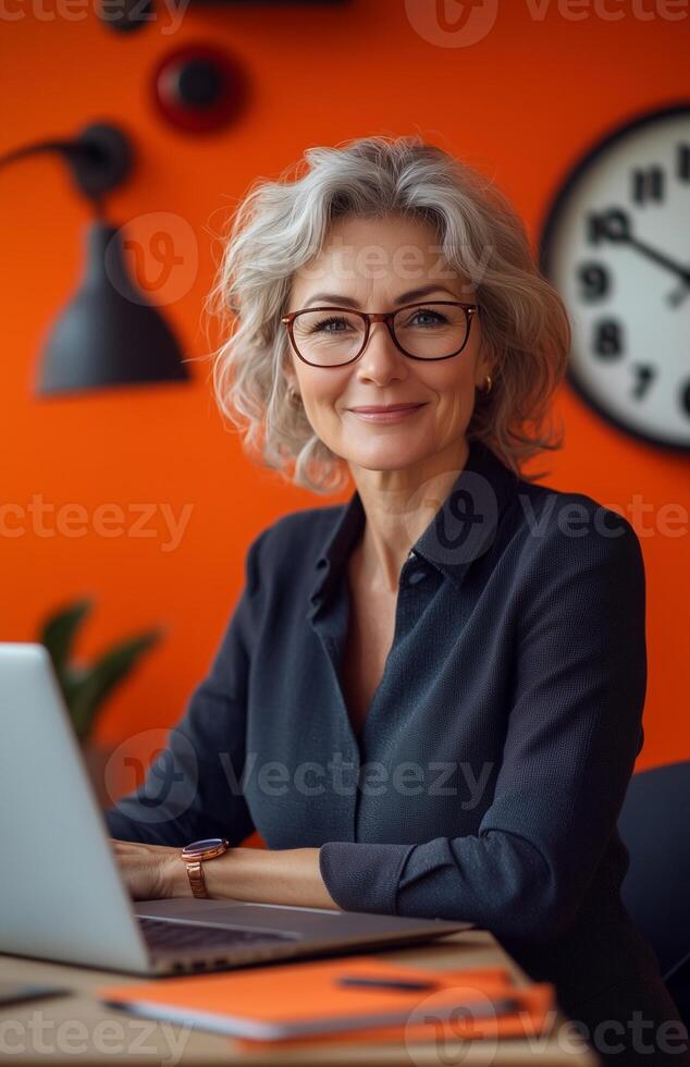 A smiling middle aged woman in an orange office, seated at her desk with a laptop photo