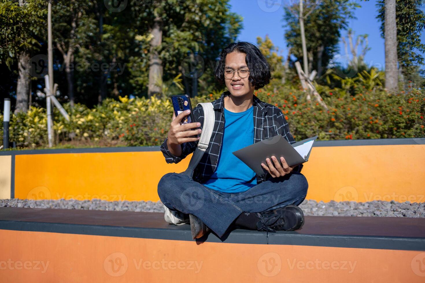 A male student wearing a checked shirt and glasses sits in the park taking a selfie with his phone. photo