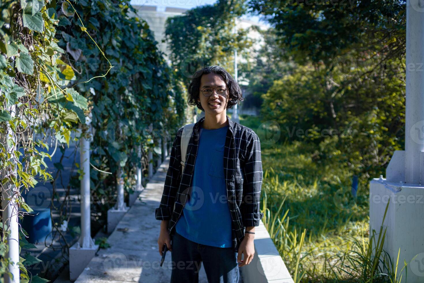 A male student wearing a plaid shirt stands in the park. photo