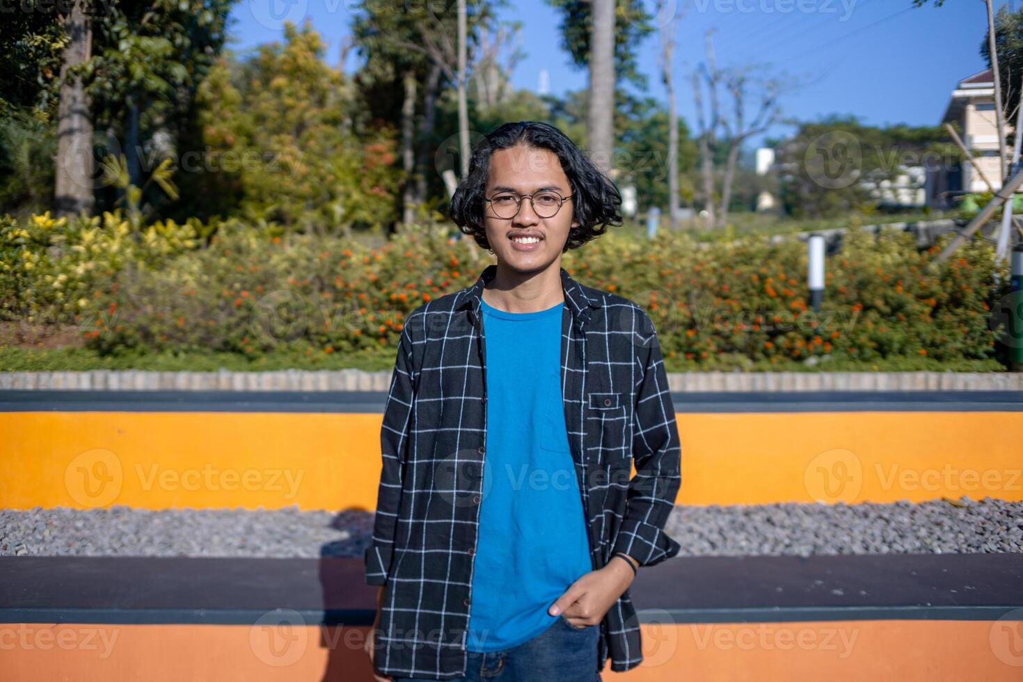 A man wearing a plaid shirt stands in the park casually. photo