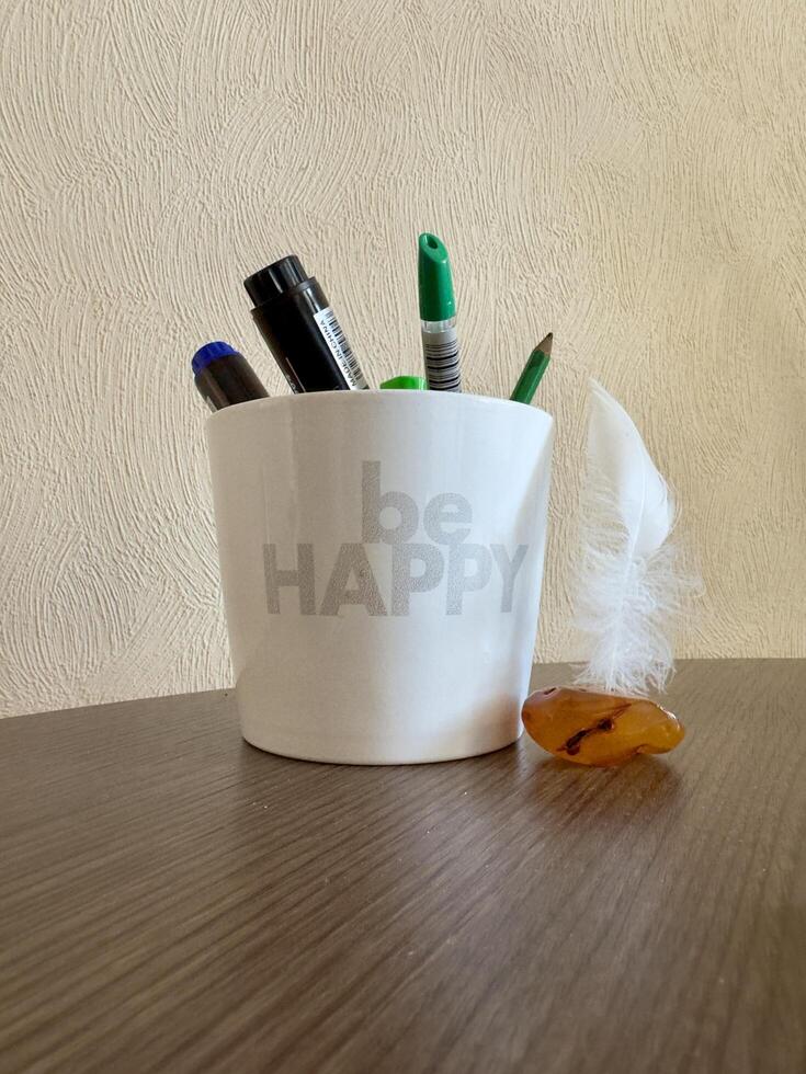 A white mug with the text Be Happy filled with pens and a marker, next to a feather and an amber stone on a wooden surface. photo
