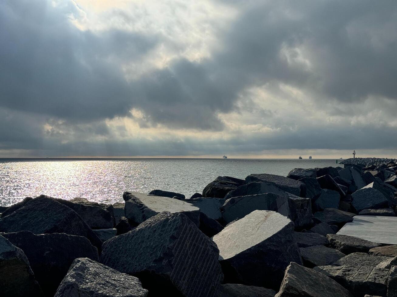 A serene seascape with large rocks in the foreground, distant ships preparing to enter the harbor, under a dramatic cloudy sky with sunbeams shining through. photo