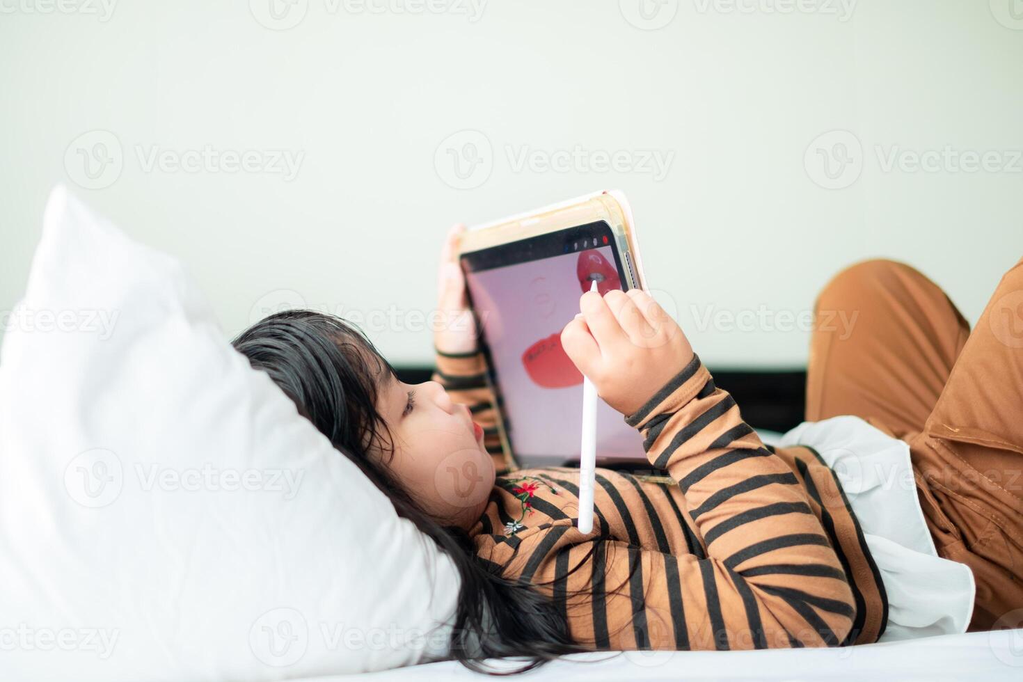 Cute Asian girl, 5-6 years old, sitting and playing with a tablet on a soft white mattress, online education learning technology, early childhood education, internet learning. photo