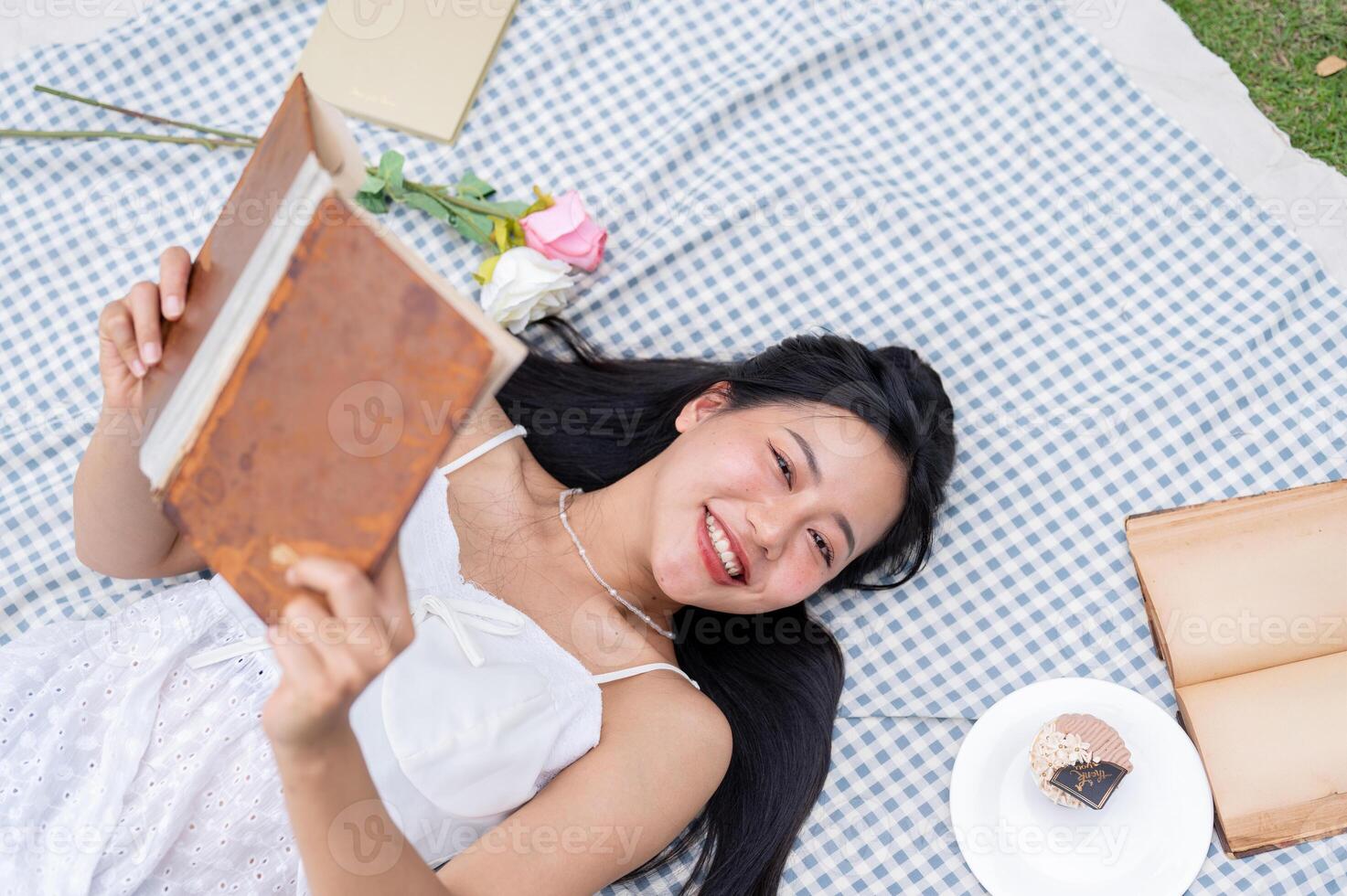 A top view of a positive, lovely Asian woman reading a book and lying on a picnic blanket. photo