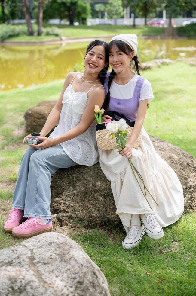Two lovely Asian woman in cute dresses are sitting on a stone together in a park. photo