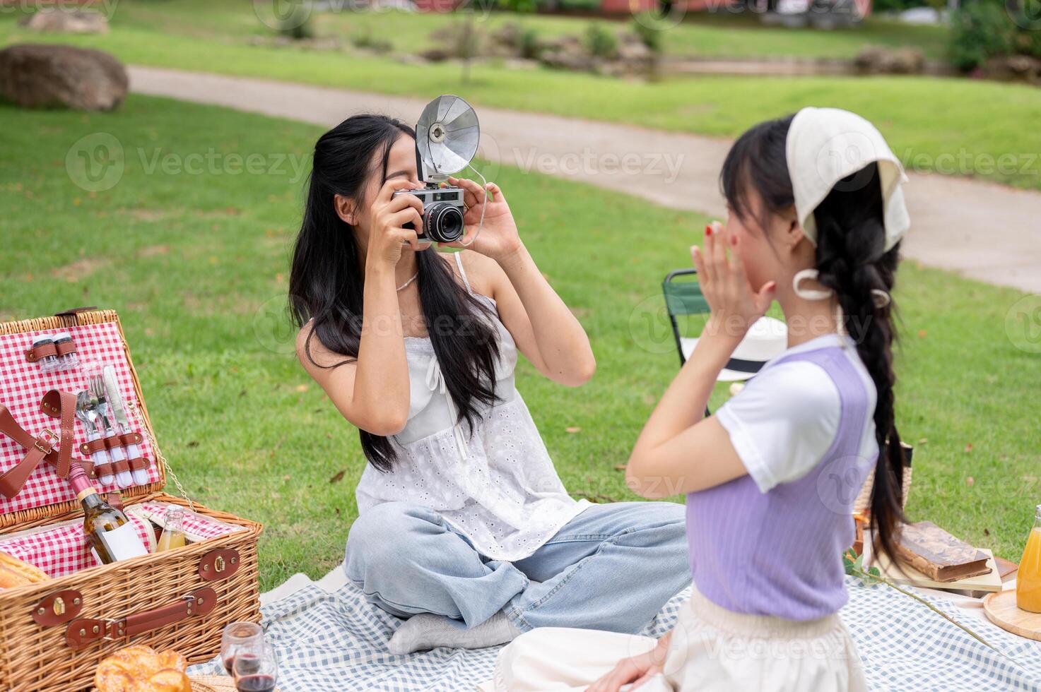 A happy Asian woman takes a picture of her friend with a retro camera while they enjoy a picnic. photo