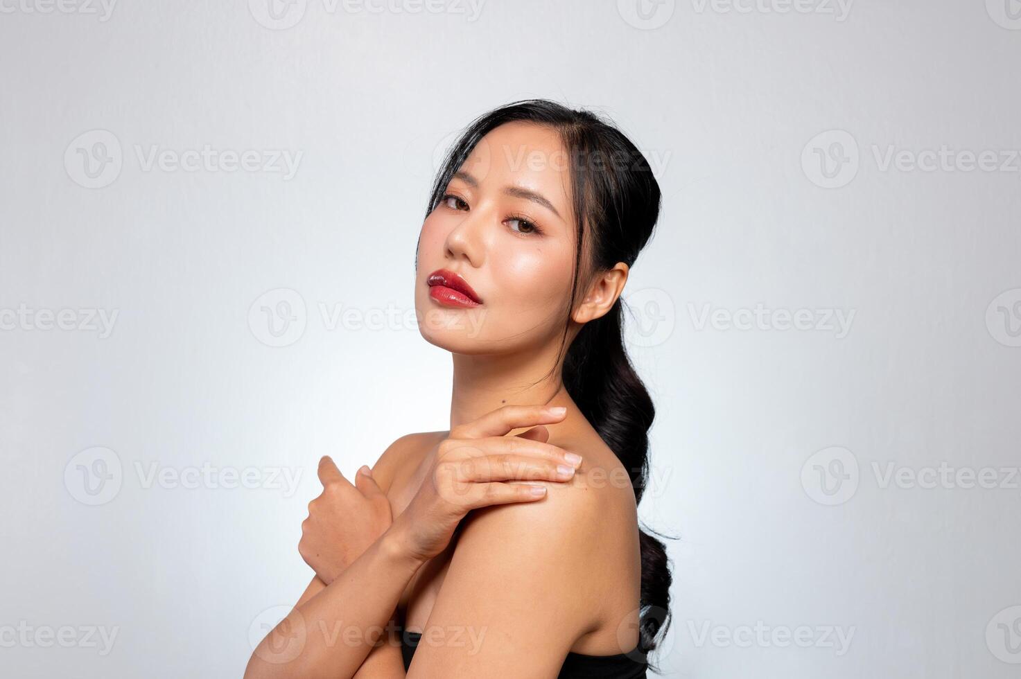 A gorgeous Asian model touches her glowing tanned skin while posing against a grey studio background photo
