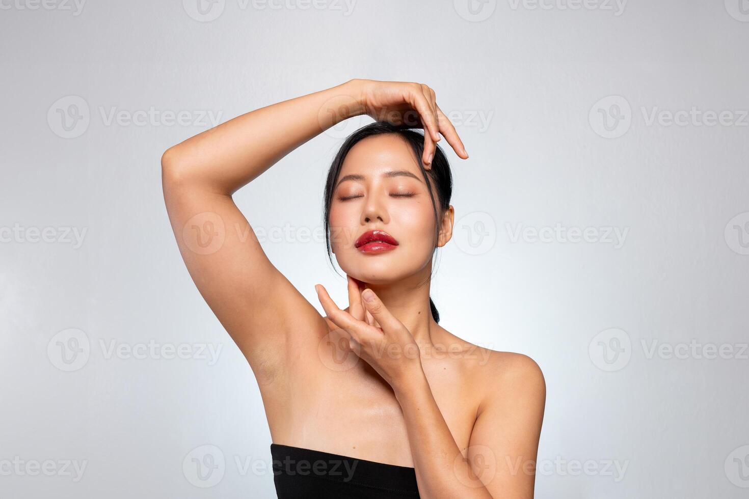 A beautiful Asian woman poses against a grey background, touching her face with her eyes closed. photo