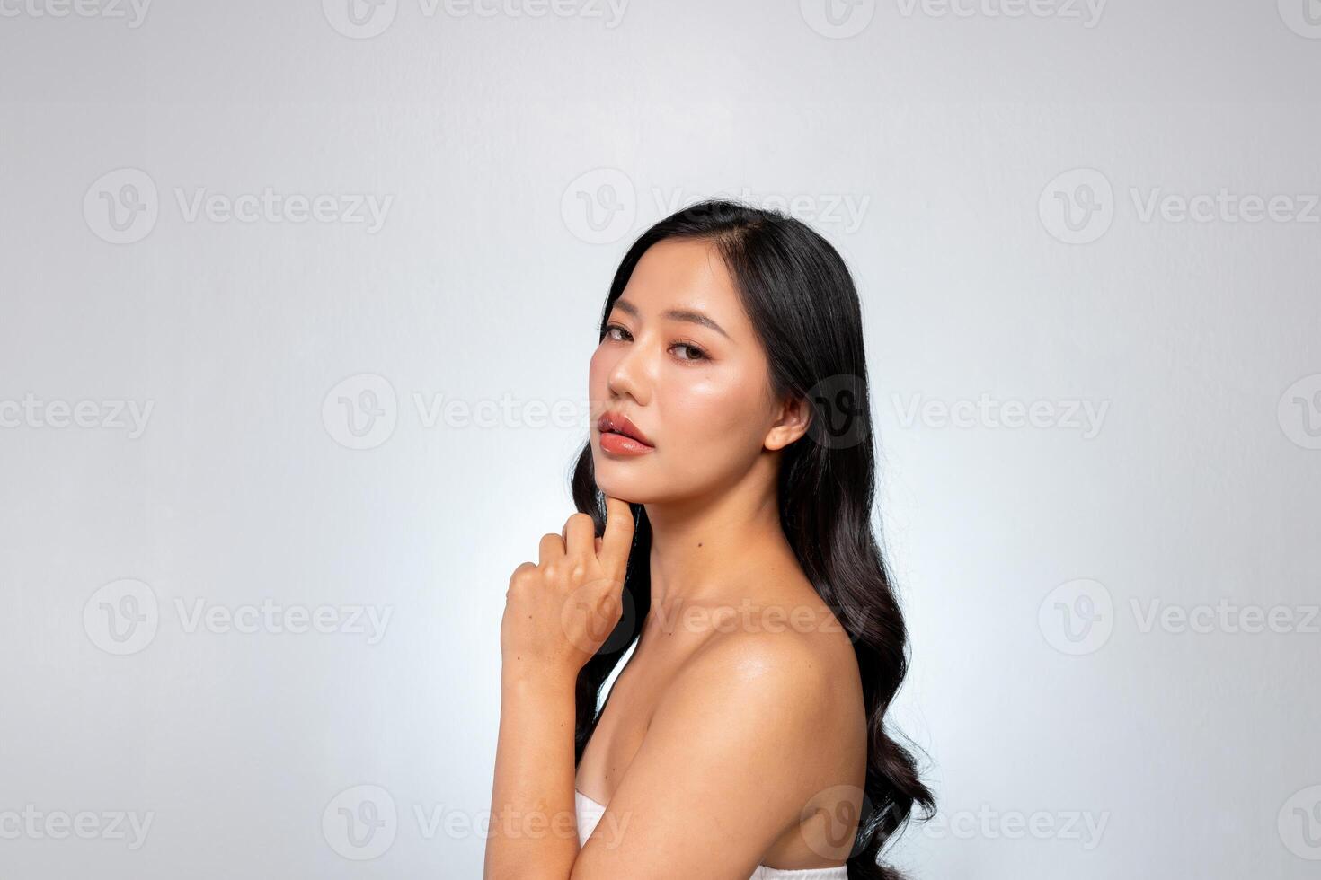 Elegant Asian woman poses against a grey background, looking at the camera while touching her chin. photo