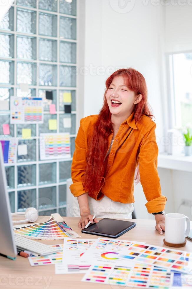 Cheerful Asian female graphic designer is standing leaning against the table, working in the studio. photo
