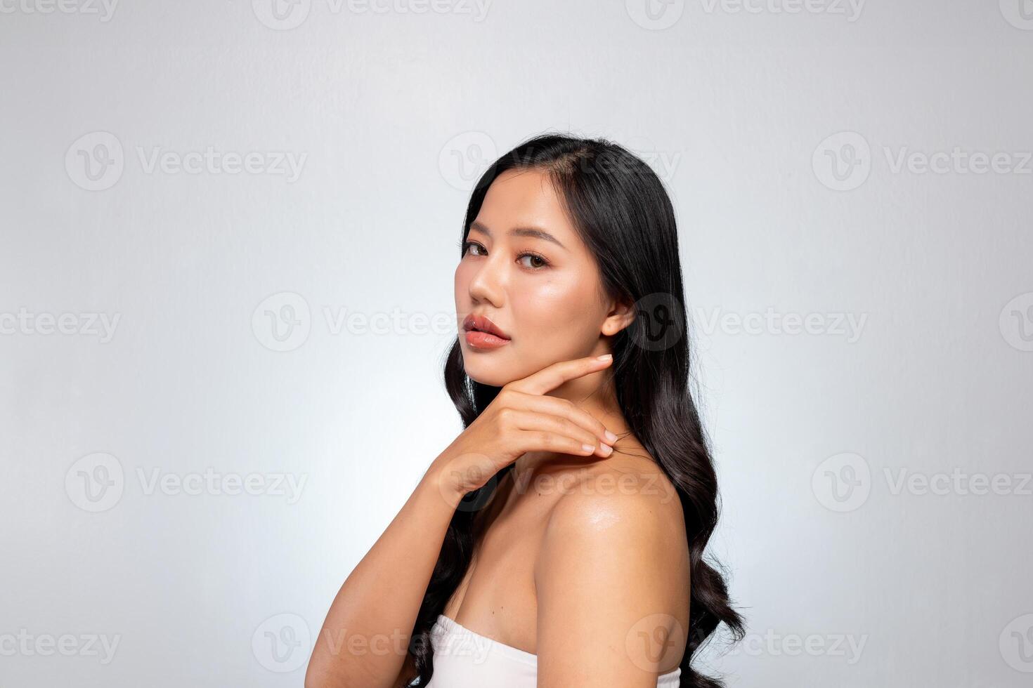 Elegant Asian woman poses against a grey background, looking at the camera while touching her chin. photo