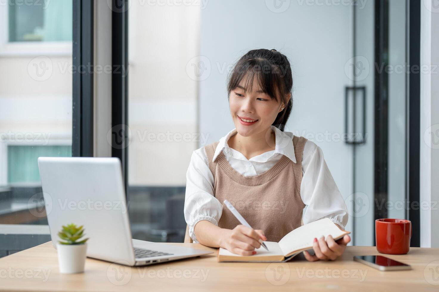 An Asian female college student is lecturing in a book while studying an online class via her laptop photo