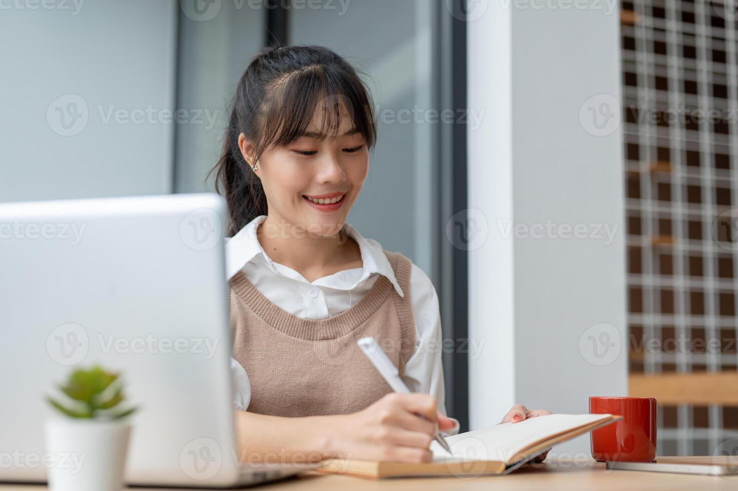 An Asian female college student is lecturing in a book while studying an online class via her laptop photo