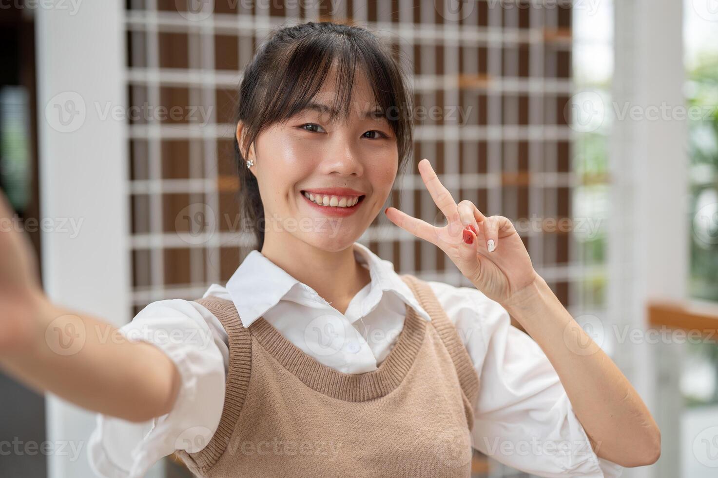 An attractive Asian woman is showing a two-fingers pose and smiling at the camera, taking a selfie. photo
