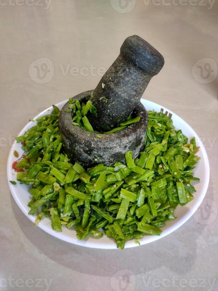 A stone mortar and pestle filled with finely chopped green cluster beans rests on a white plate. photo