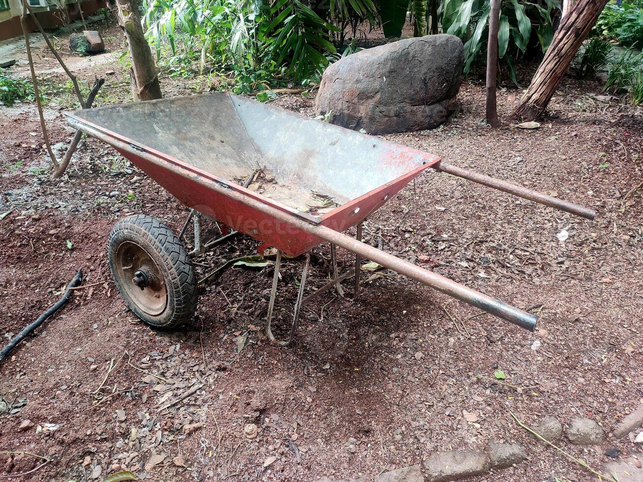 A Red wheelbarrow sitting outdoors on a patch of dirt with some leaves scattered around. There's a large rock and some greenery in the background. photo