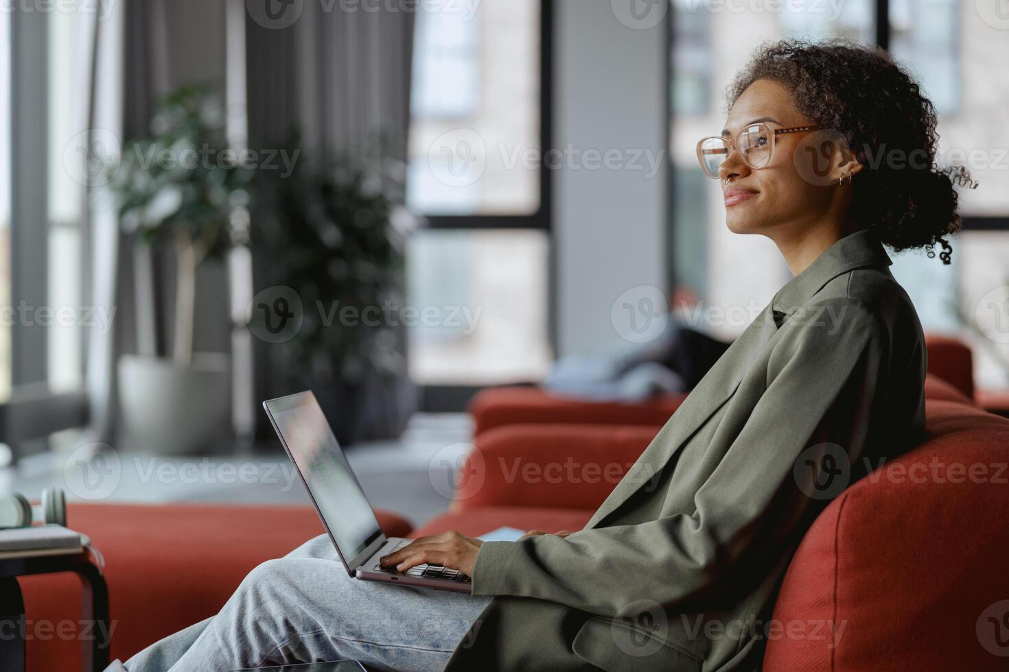 Woman on couch using laptop computer in comfort near window with houseplant in nice modern coworking office photo