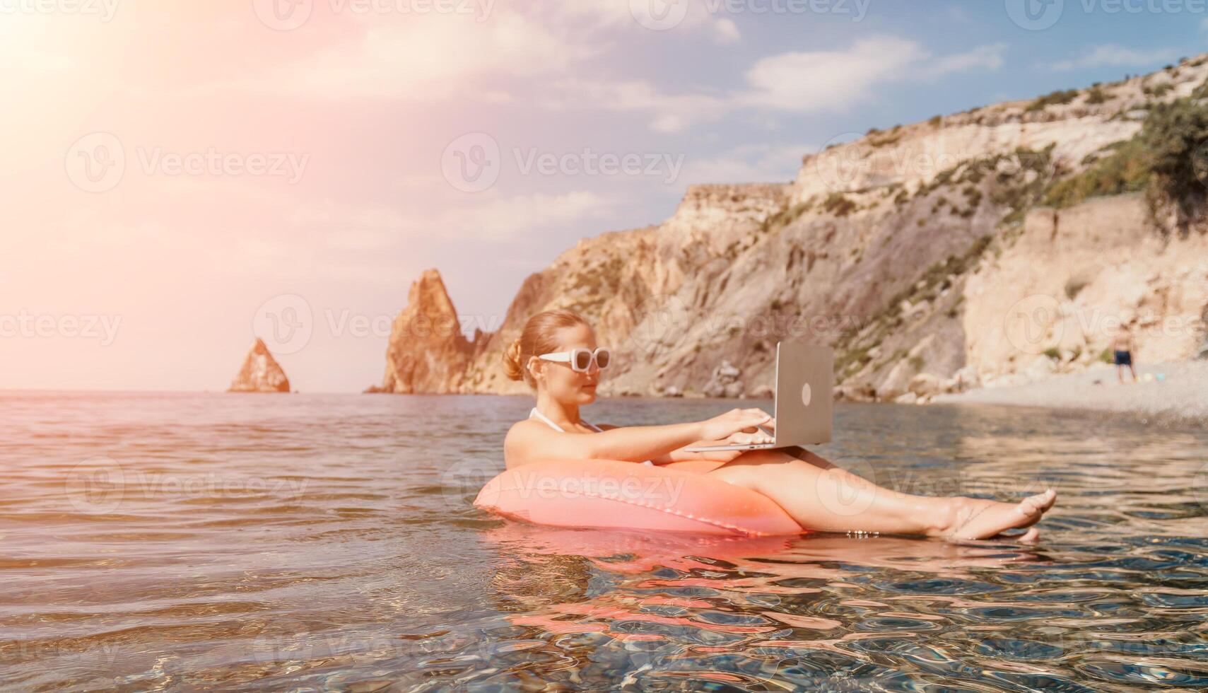 Woman freelancer works on laptop swimming in sea on pink inflatable ring. Happy tourist in sunglasses floating on inflatable donut and working on laptop computer in calm ocean. Remote working anywhere photo