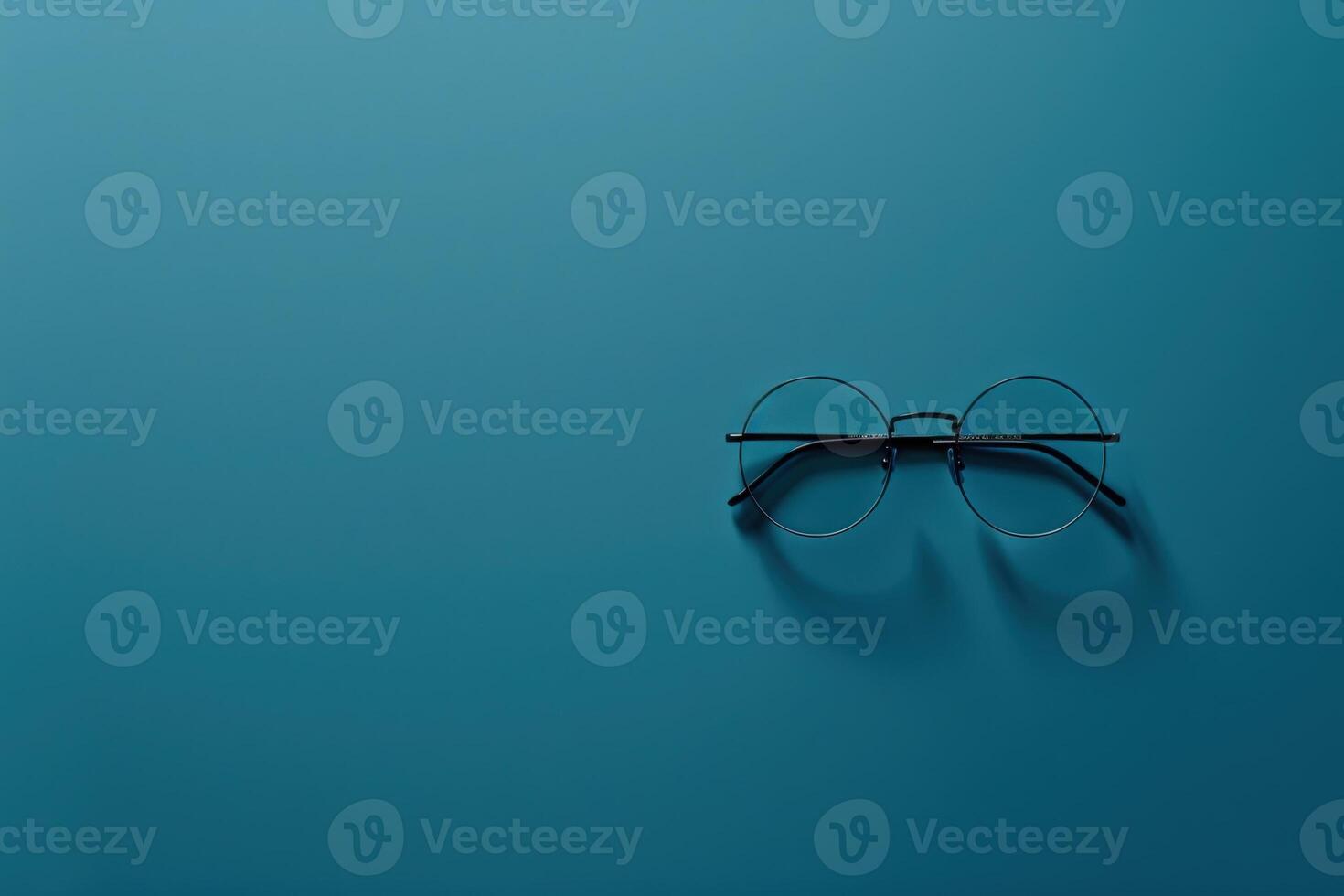 A pair of fashionable glasses lies alone on a deep teal surface, casting delicate shadows while inviting exploration. photo