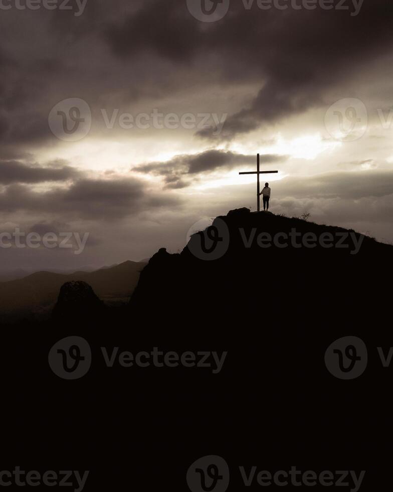 Silhouette of hopeful woman standing alone by cross search for purpose on scenic hilltop viewpoint with dramatic sky background sunshine on sunset.Conceptual loneliness, despair, search for purpose photo