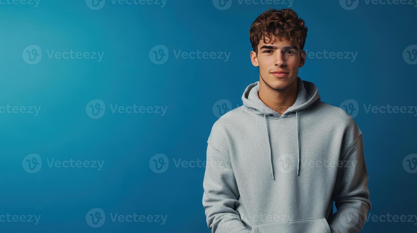Young man wearing a gray hoodie stands against a blue background, looking confidently at the camera in a studio setting photo
