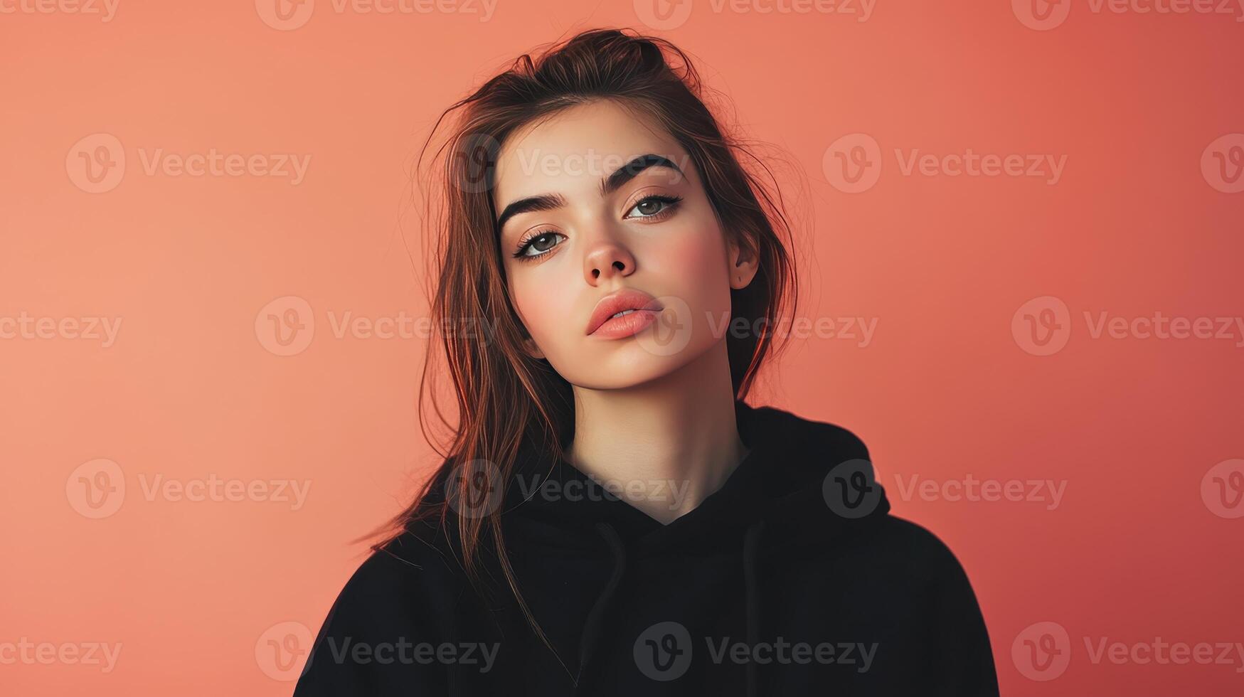 Young woman with natural makeup posing against a peach-colored backdrop in a casual black hoodie during daylight photo