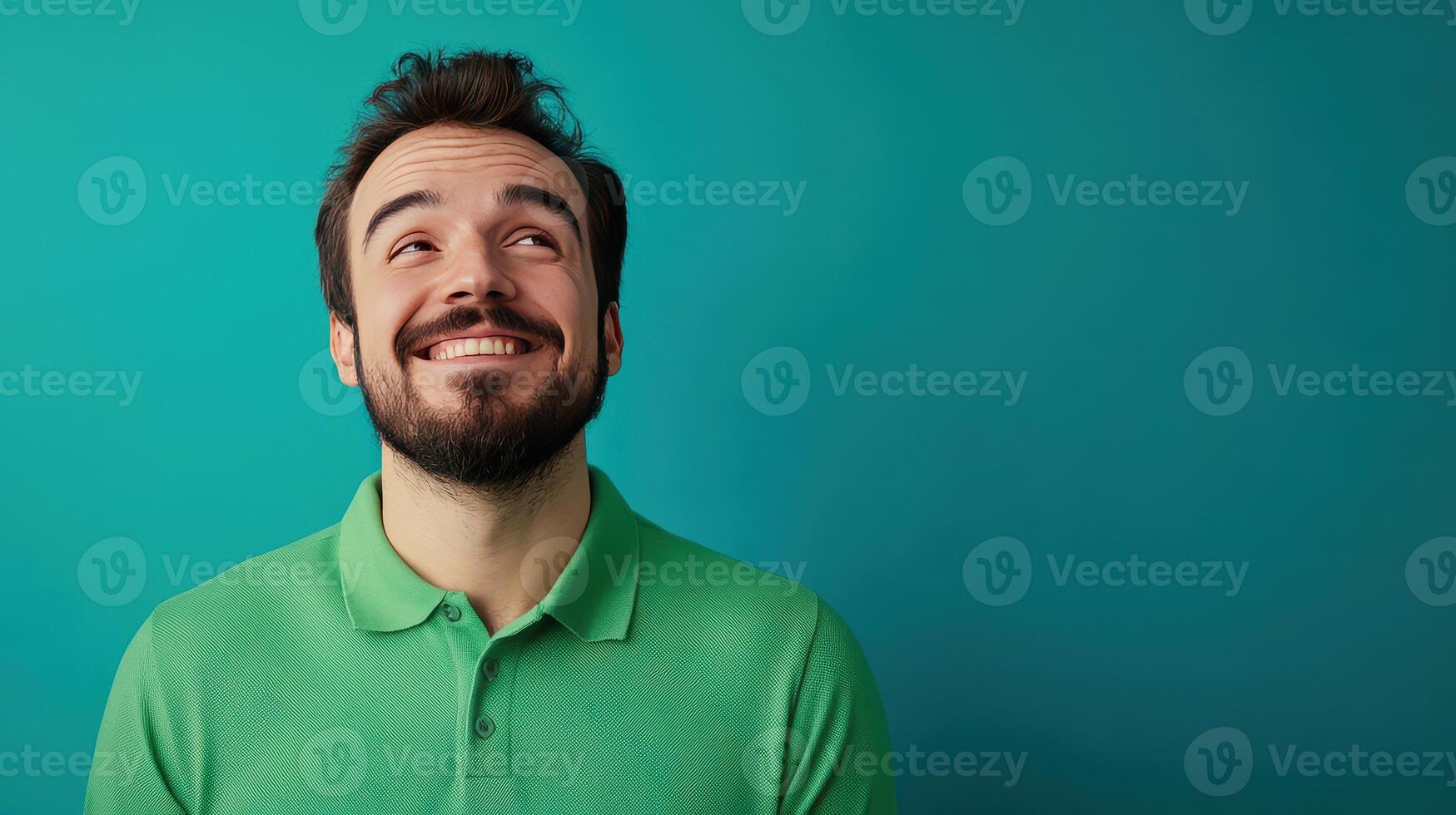 Smiling man in a green polo shirt against a solid teal background, looking upwards with a cheerful expression photo