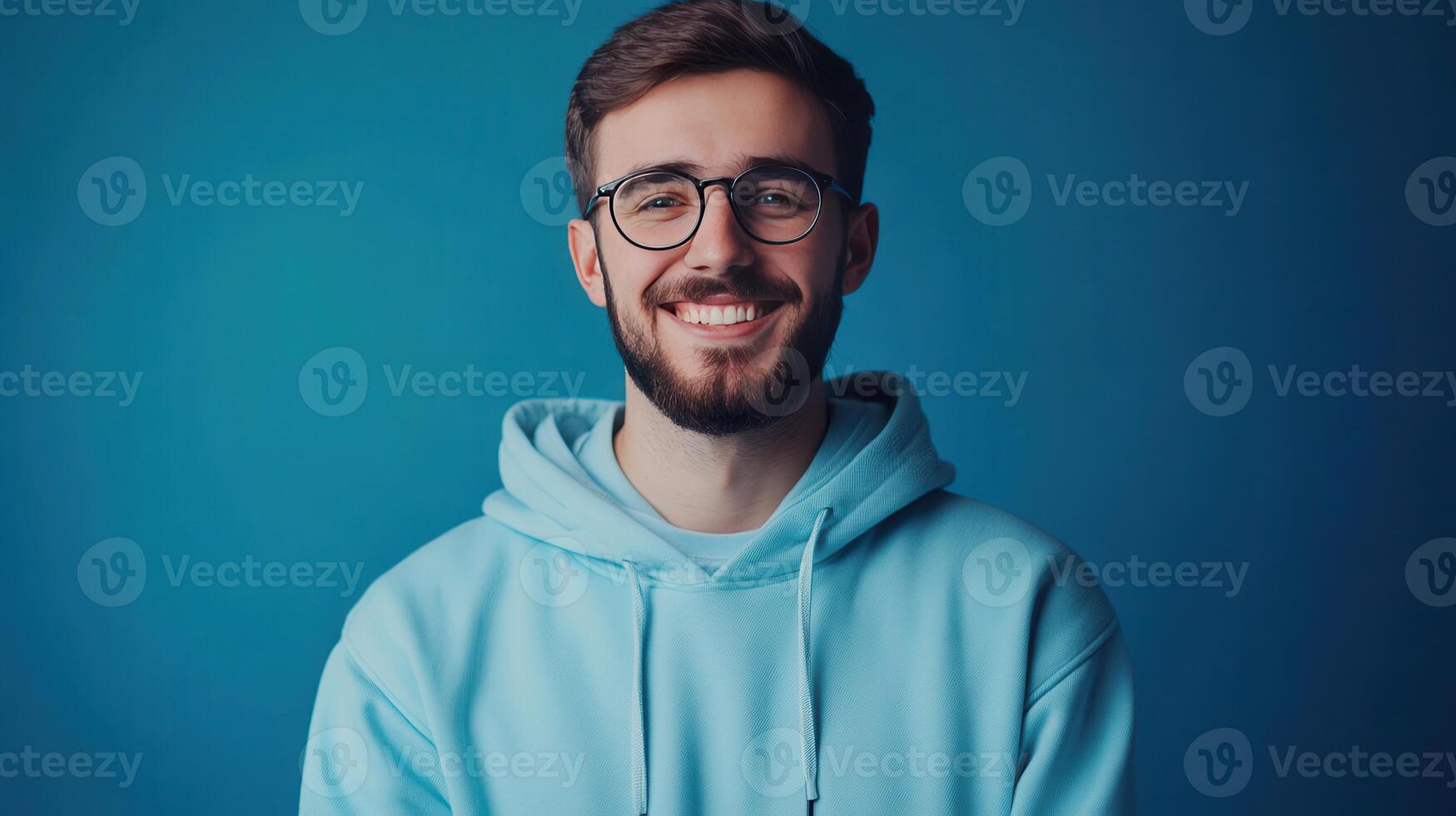 Young man smiling warmly while wearing a light blue hoodie against a solid blue background in a relaxed indoor setting photo