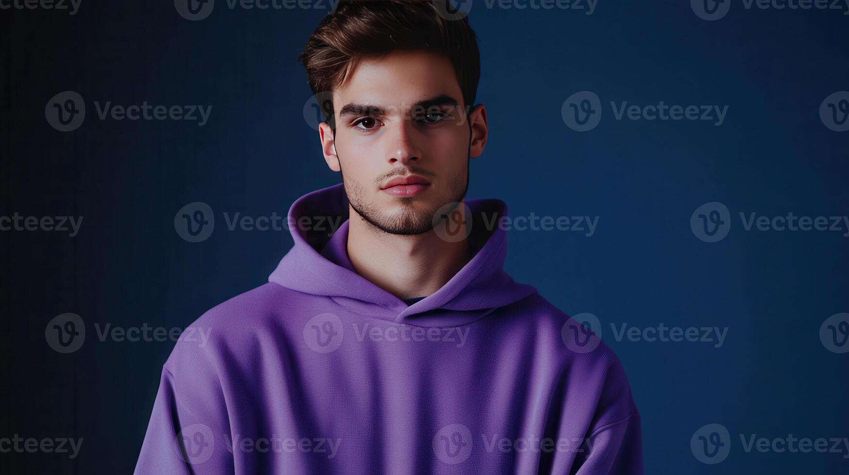 Young man in a purple hoodie stands against a dark blue background, looking thoughtfully into the camera photo