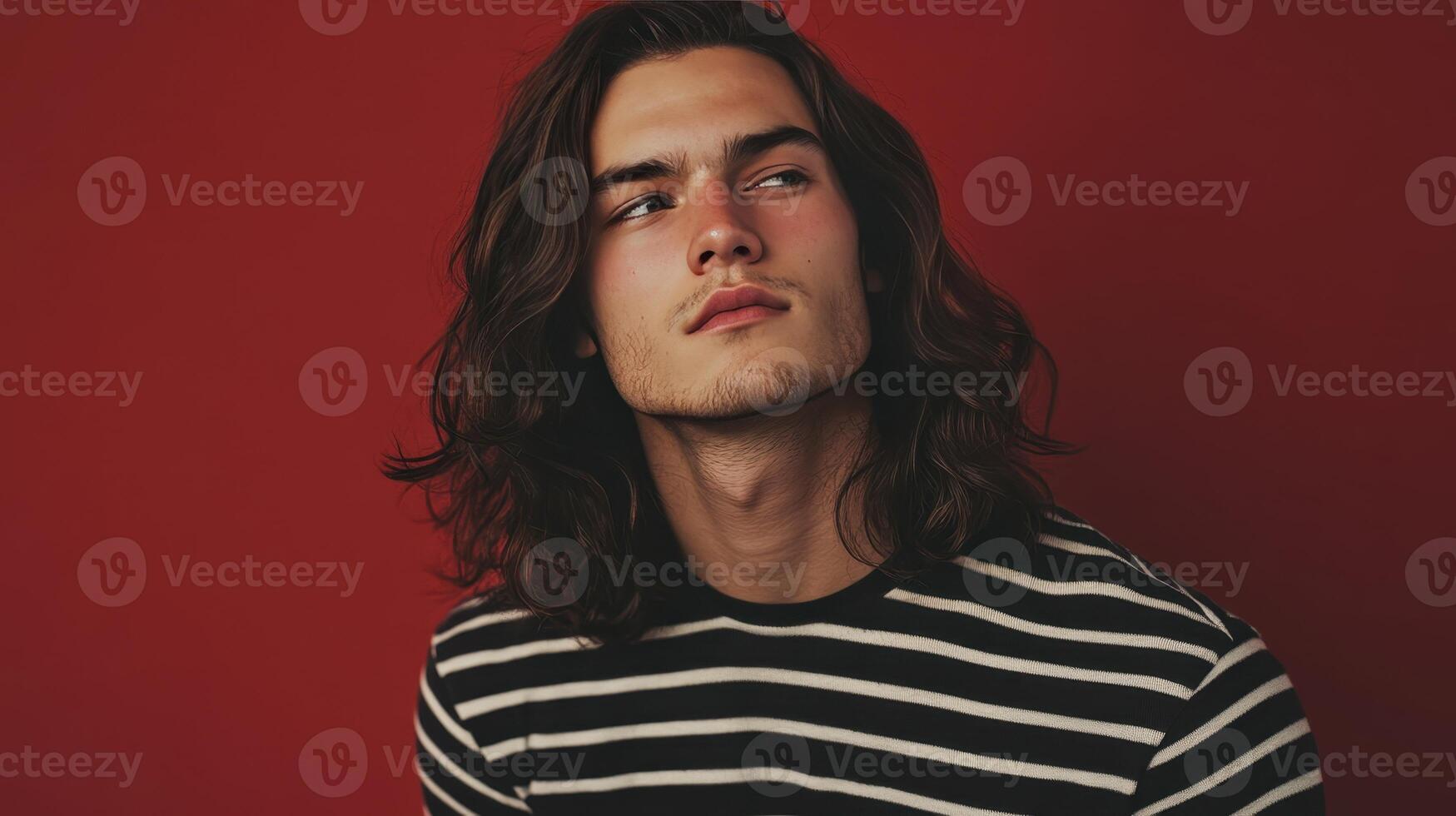 Young man with long hair wearing a striped shirt poses against a red wall in a studio setting during golden hour photo