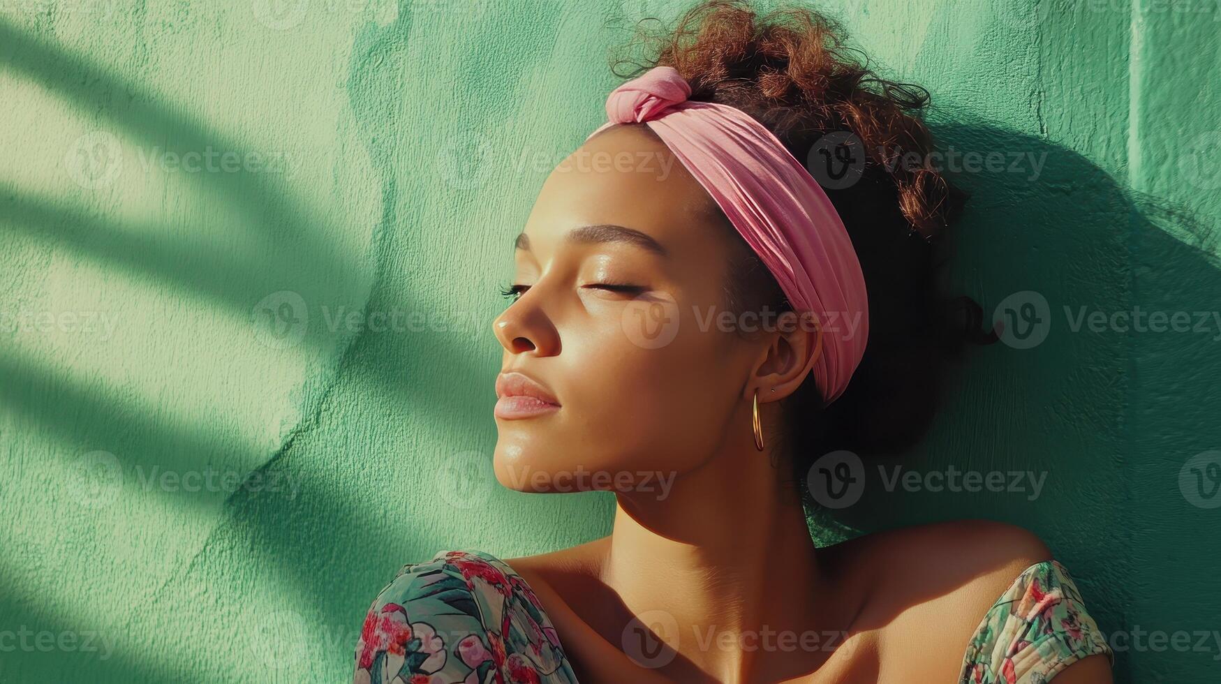 Woman relaxing against a turquoise wall while enjoying sunlight, wearing a floral top and a pink headband photo