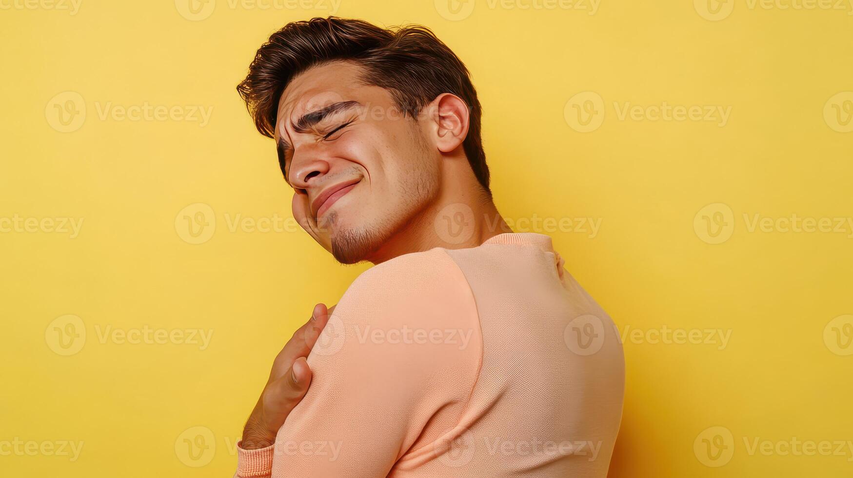 Young man smiles playfully against a bright yellow background while hugging himself in a light sweater photo