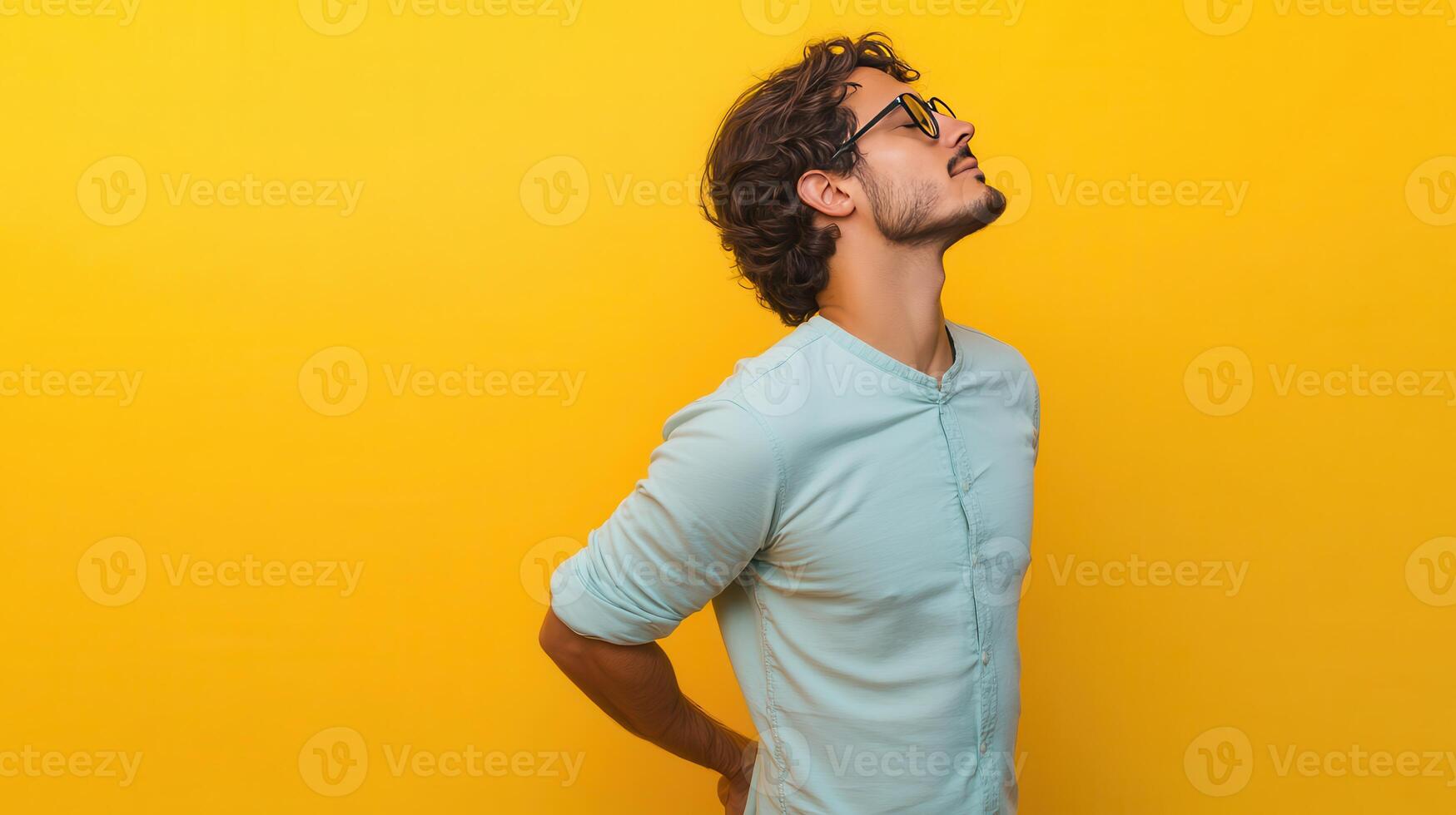 Young man looking upwards against a bright yellow background with hands behind his back indoors photo