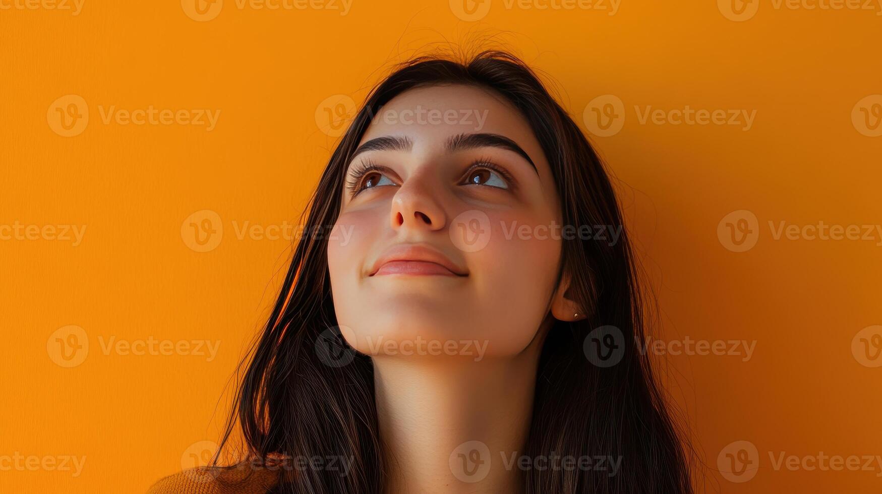 Young woman gazing upward against a vibrant orange background, expressing curiosity and contemplation in a bright indoor setting photo