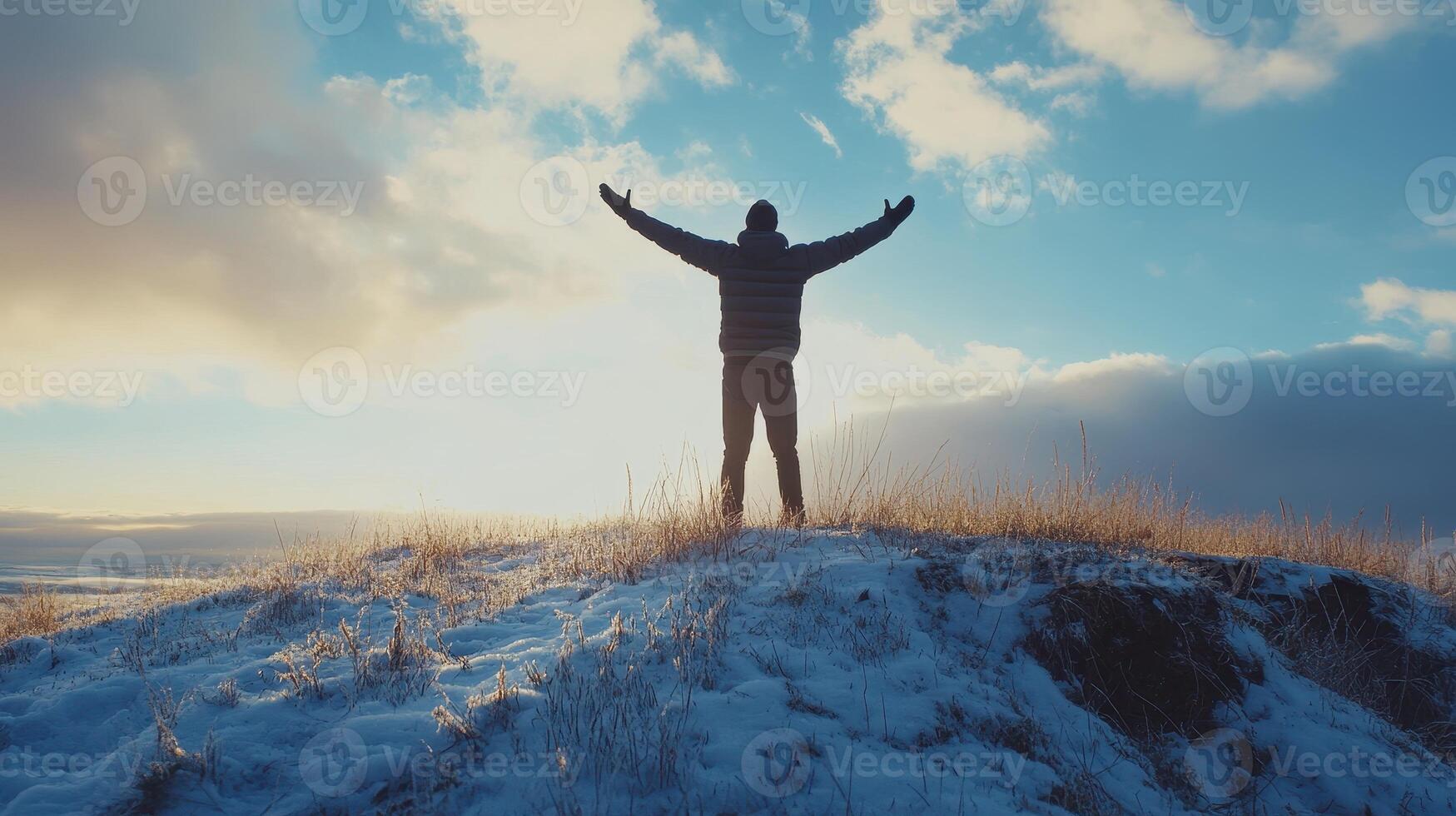 Person standing on a snowy hilltop with arms raised against a bright sky at sunset photo