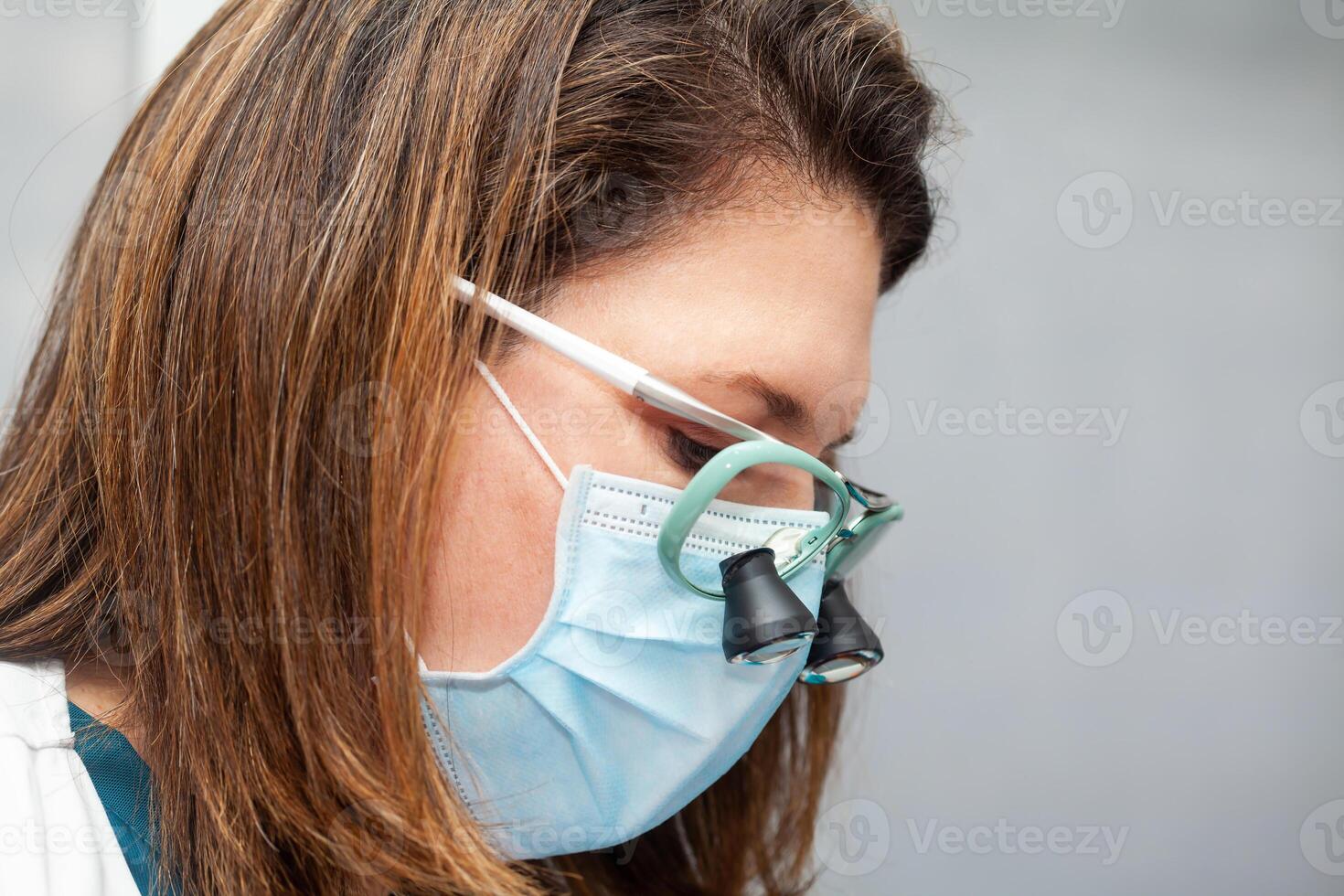 Close up of a female doctor face while suturing a patient during a small surgery. Dermatologist carrying out a removal of benign skin lesion on the face of a young male patient. photo