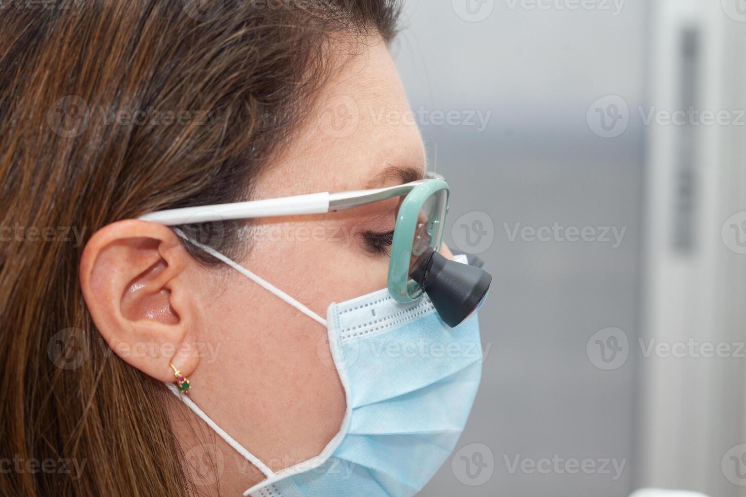 Close up of a female dermatologist face while suturing a patient during a small surgery wearing a binocular loupe. photo