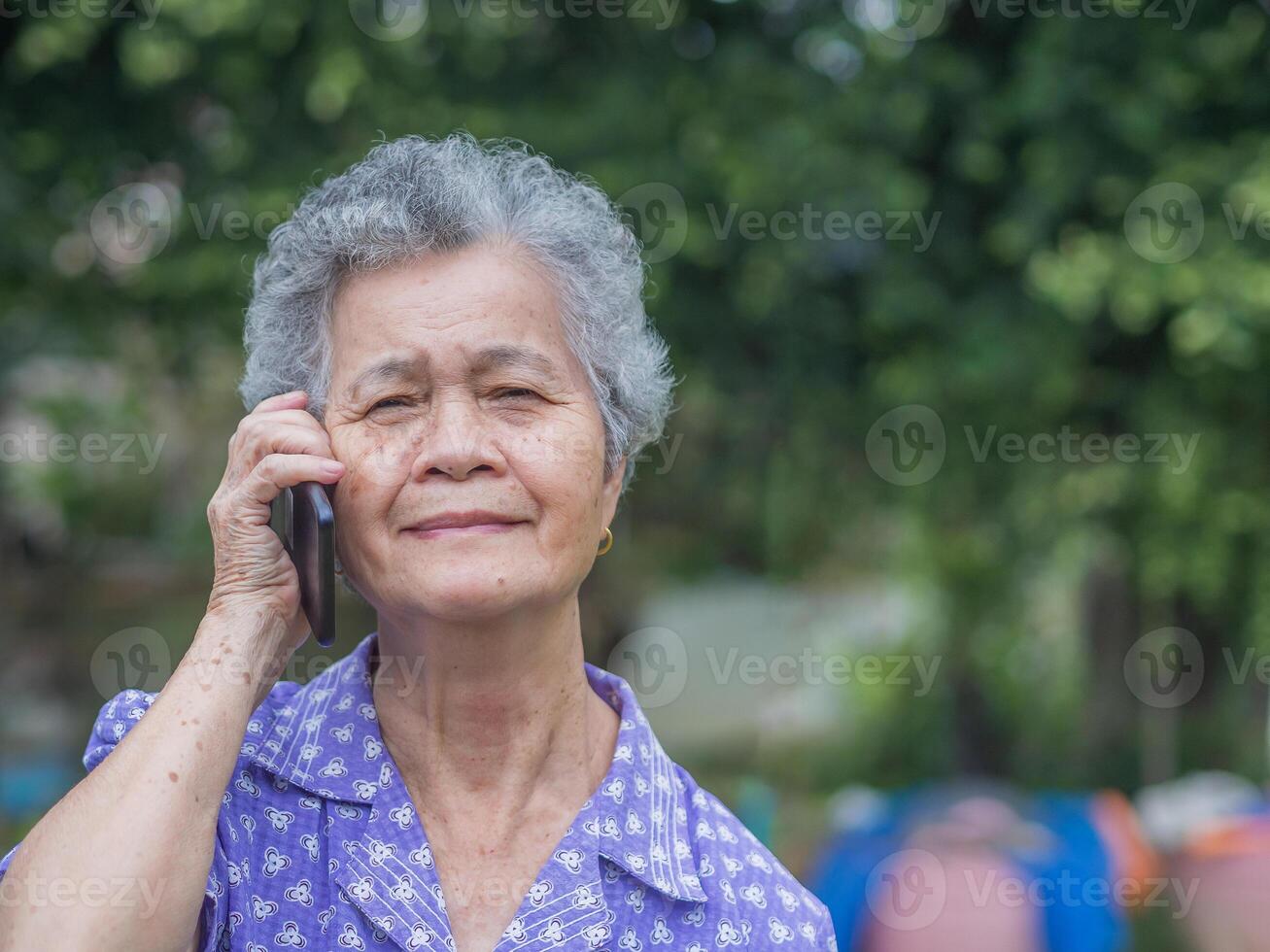 Happy beautiful senior woman is using a smartphone, smiling and looking at the camera while standing in a garden. Space for text. Aged people and communication concept photo