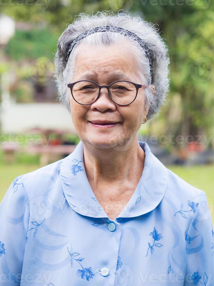 Portrait of a senior woman with short gray hair, wearing glasses, and looking at the camera with a smile while standing in a garden. Aged people and relaxation concept photo