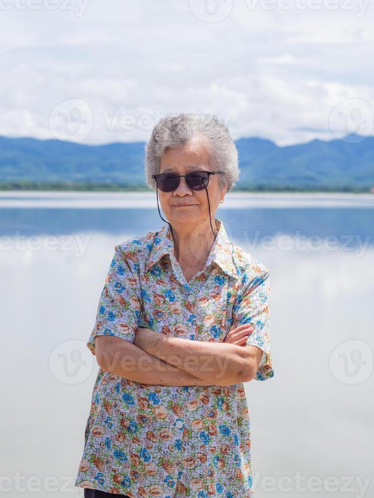 Happy a senior woman with short gray hair smiling, arms crossed, and looking at the camera standing near lake and mountains background. Concept of aged people and relaxation photo
