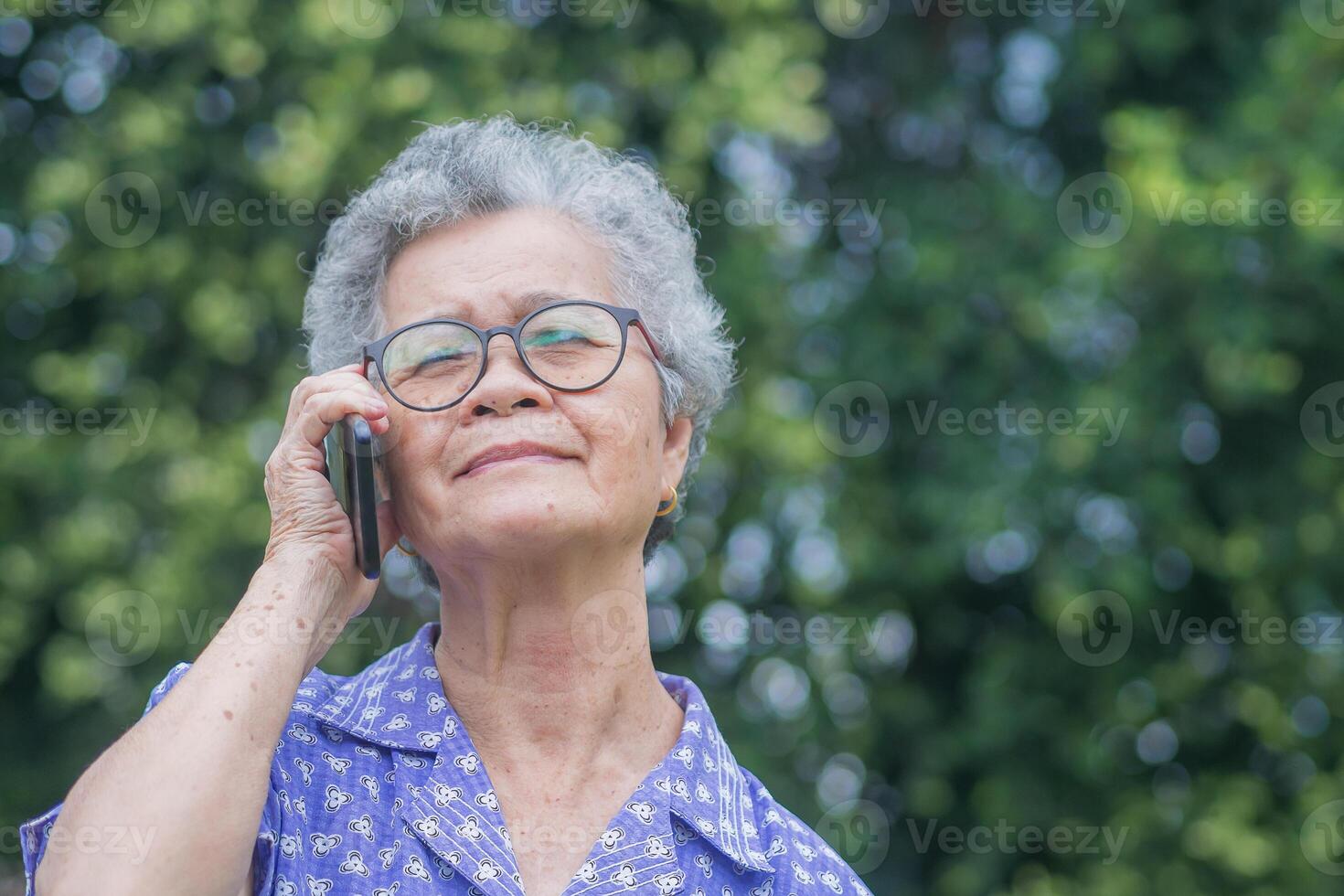 Happy beautiful a senior woman is using a smartphone, smiling and looking up while standing in a garden. Space for text. Aged people and communication concept photo