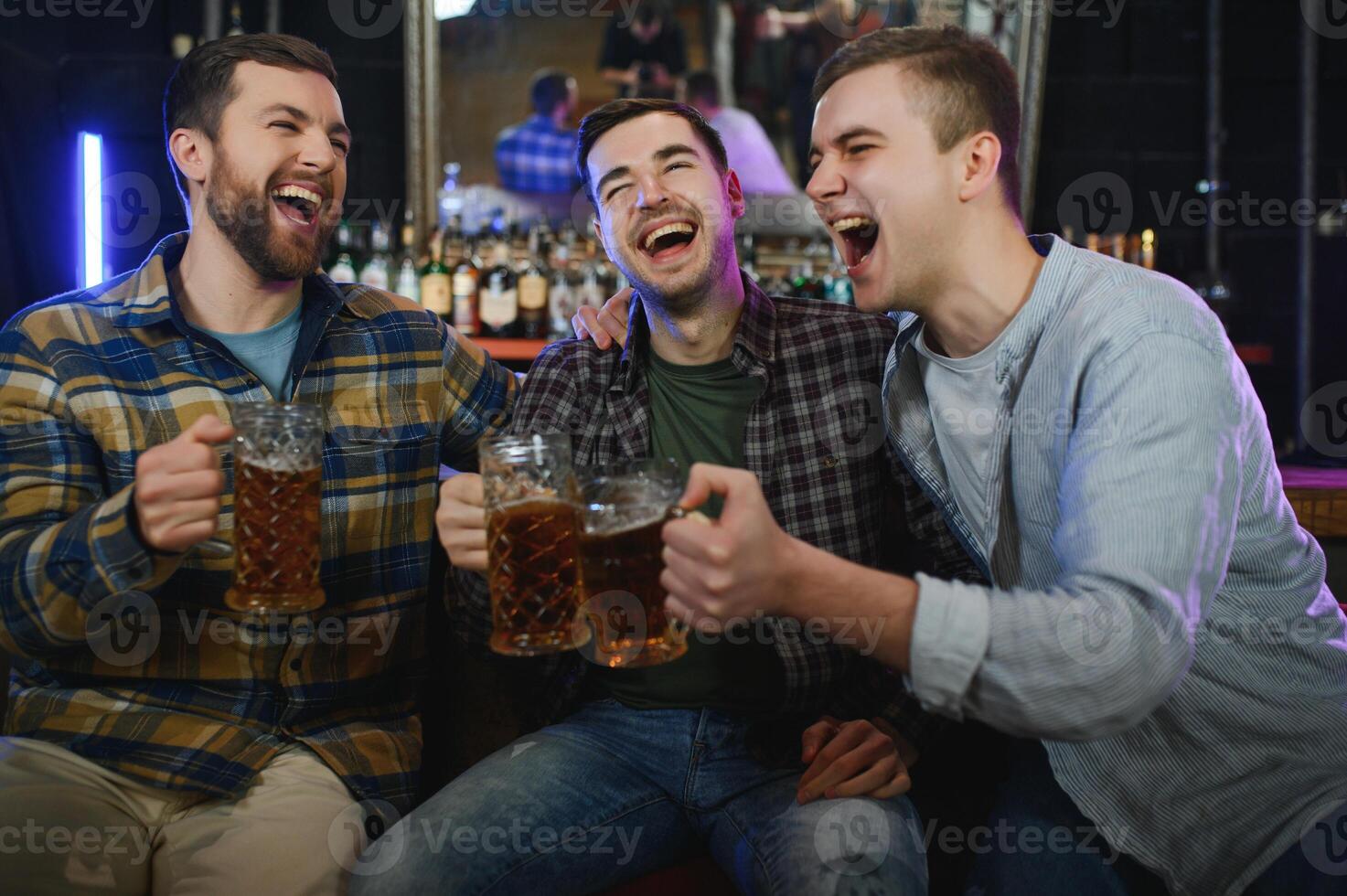 Three young men in casual clothes are smiling, holding bottles of beer while standing near bar counter in pub photo