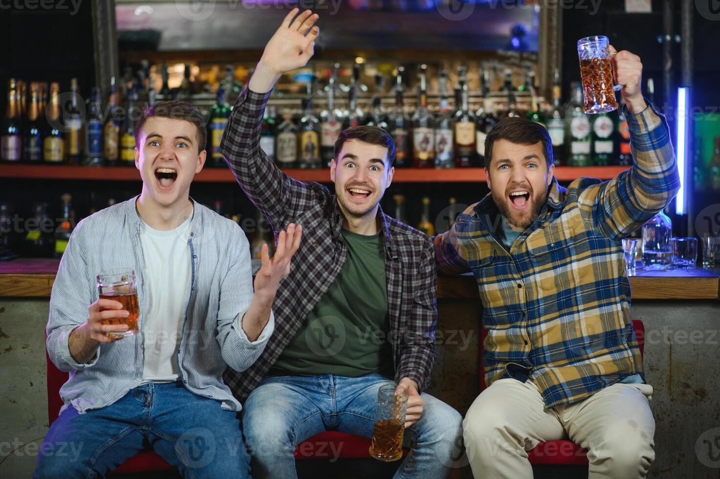 Soccer fans cheering for favourite team while enjoying drinks. Group of supporters celebrating victory while watching football match on tv in a club. Happy friends shouting while cheering for a goal. photo