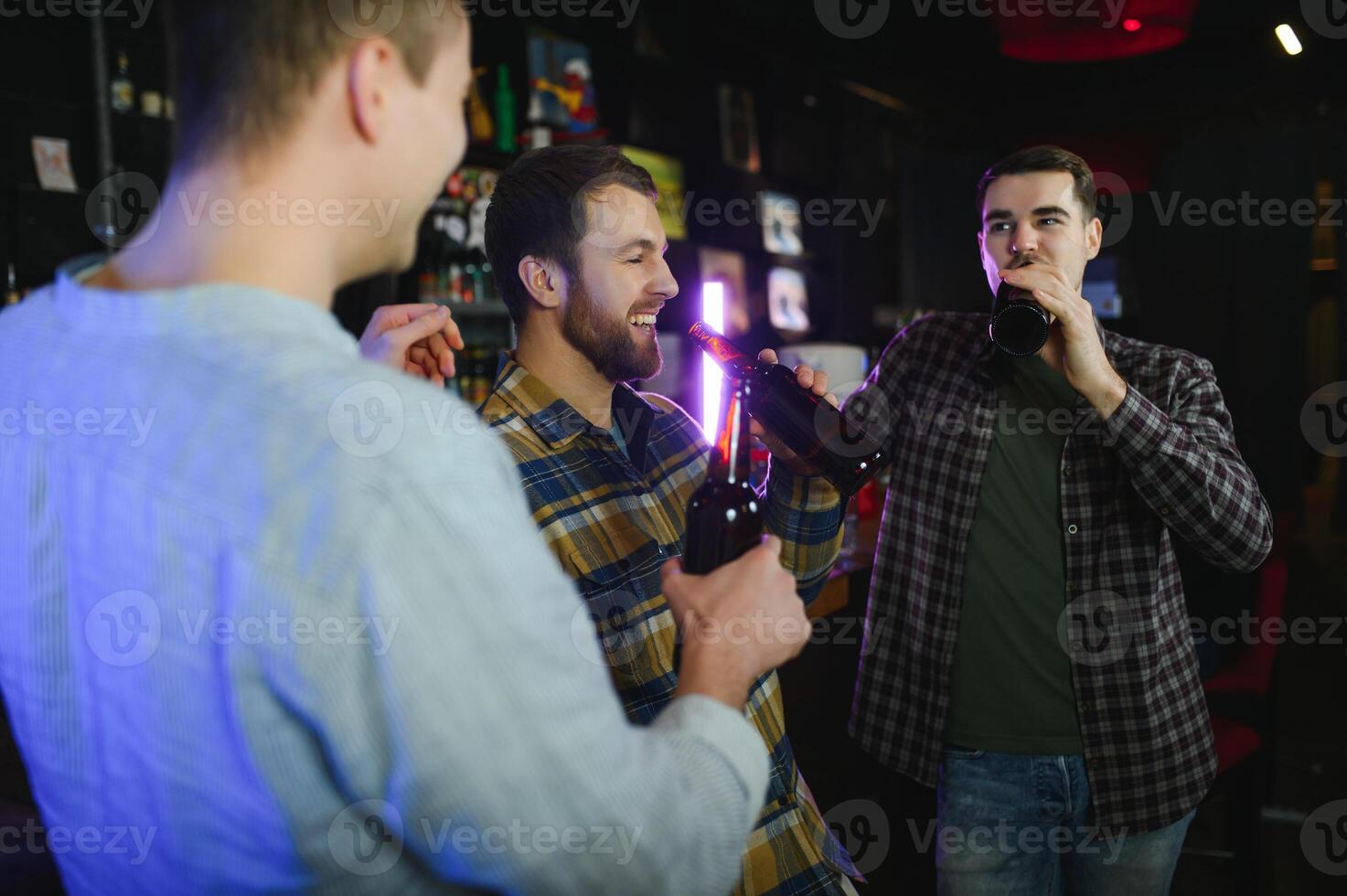 Three young men in casual clothes are smiling, holding bottles of beer while standing near bar counter in pub photo