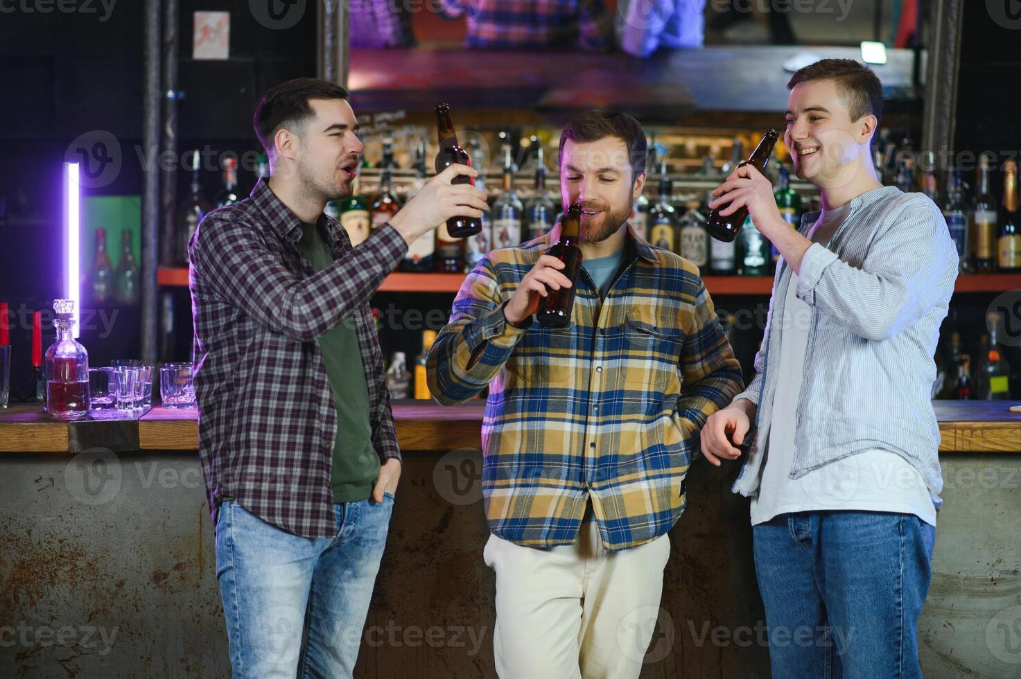 Three young men in casual clothes are smiling, holding bottles of beer while standing near bar counter in pub photo