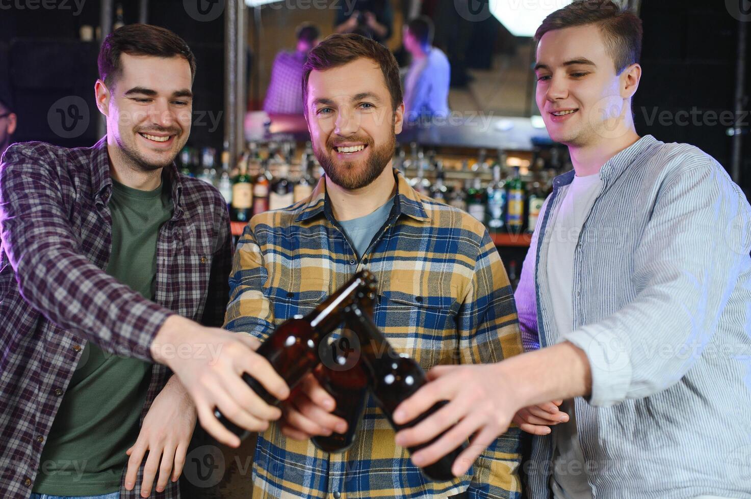 Three young men in casual clothes are smiling and clanging glasses of beer together while sitting at bar counter in pub photo
